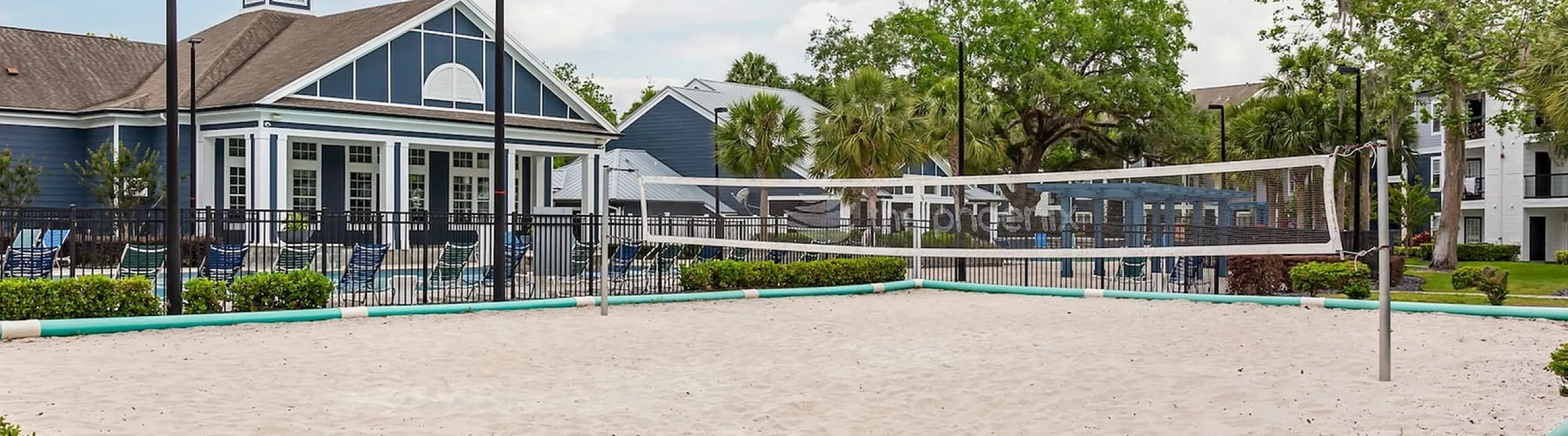 Phoenix Orlando outdoor volleyball court surrounded by lush greenery, adjacent to a modern clubhouse with comfortable lounge chairs.