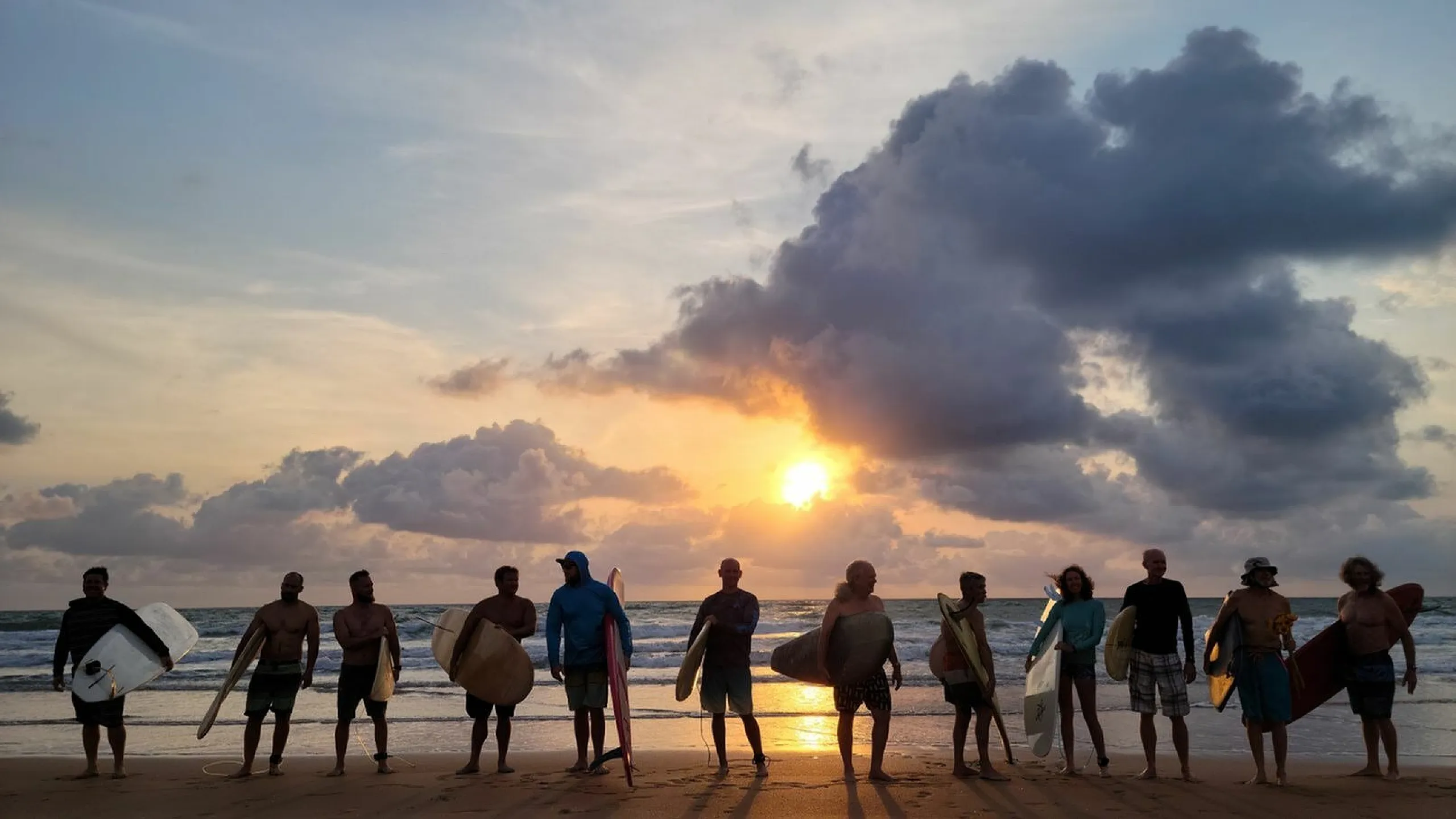 Group of surfers standing on a beach at sunset, holding surfboards and enjoying the vibrant sky and calming waves.