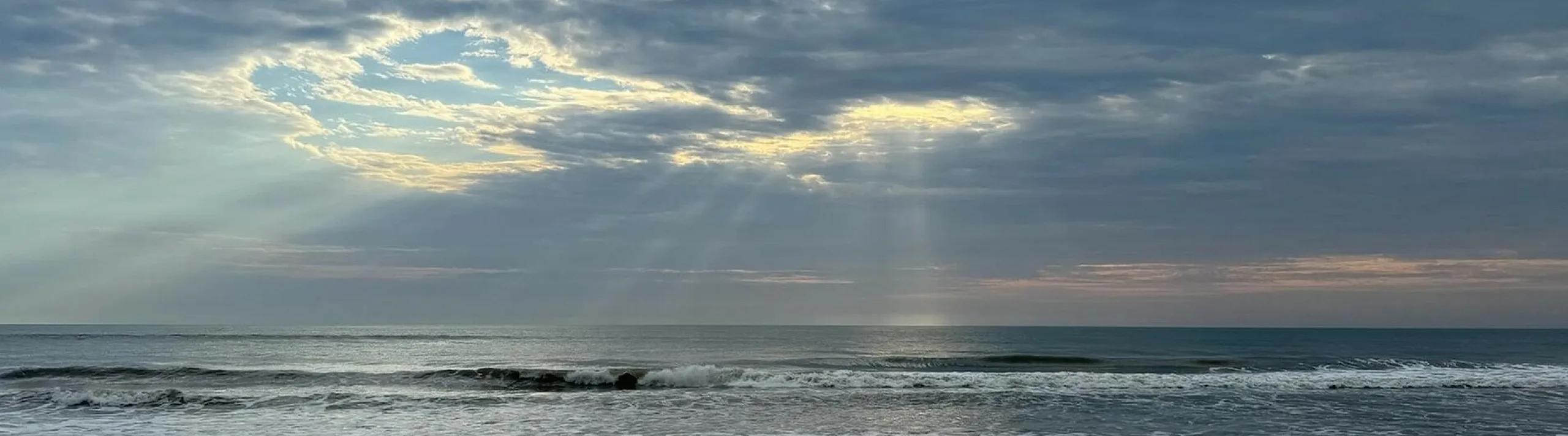 Serene beachscape with soft waves, dramatic clouds, and sunbeams illuminating the horizon, creating a peaceful coastal atmosphere.