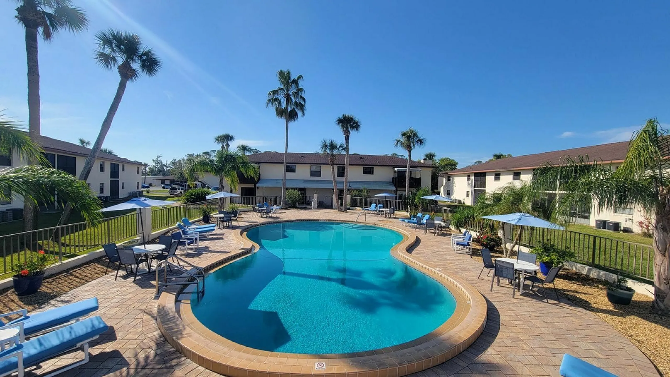 Serene apartment complex with a sparkling outdoor pool, surrounded by lush palm trees and inviting sun loungers under a bright blue sky.
