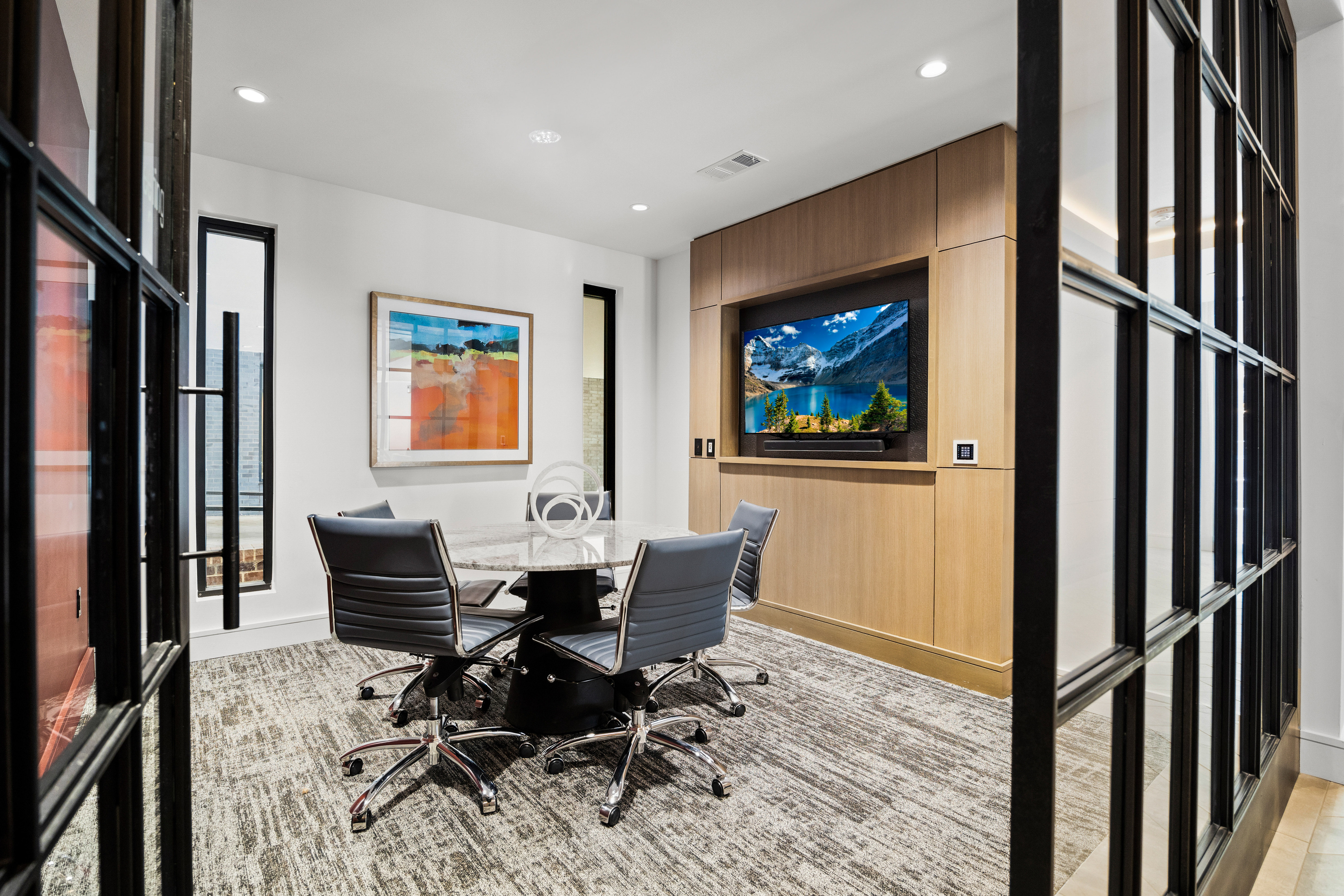 apartment community conference room with glass doors, leather chairs around a marble table, artwork, and a large wall-mounted TV.