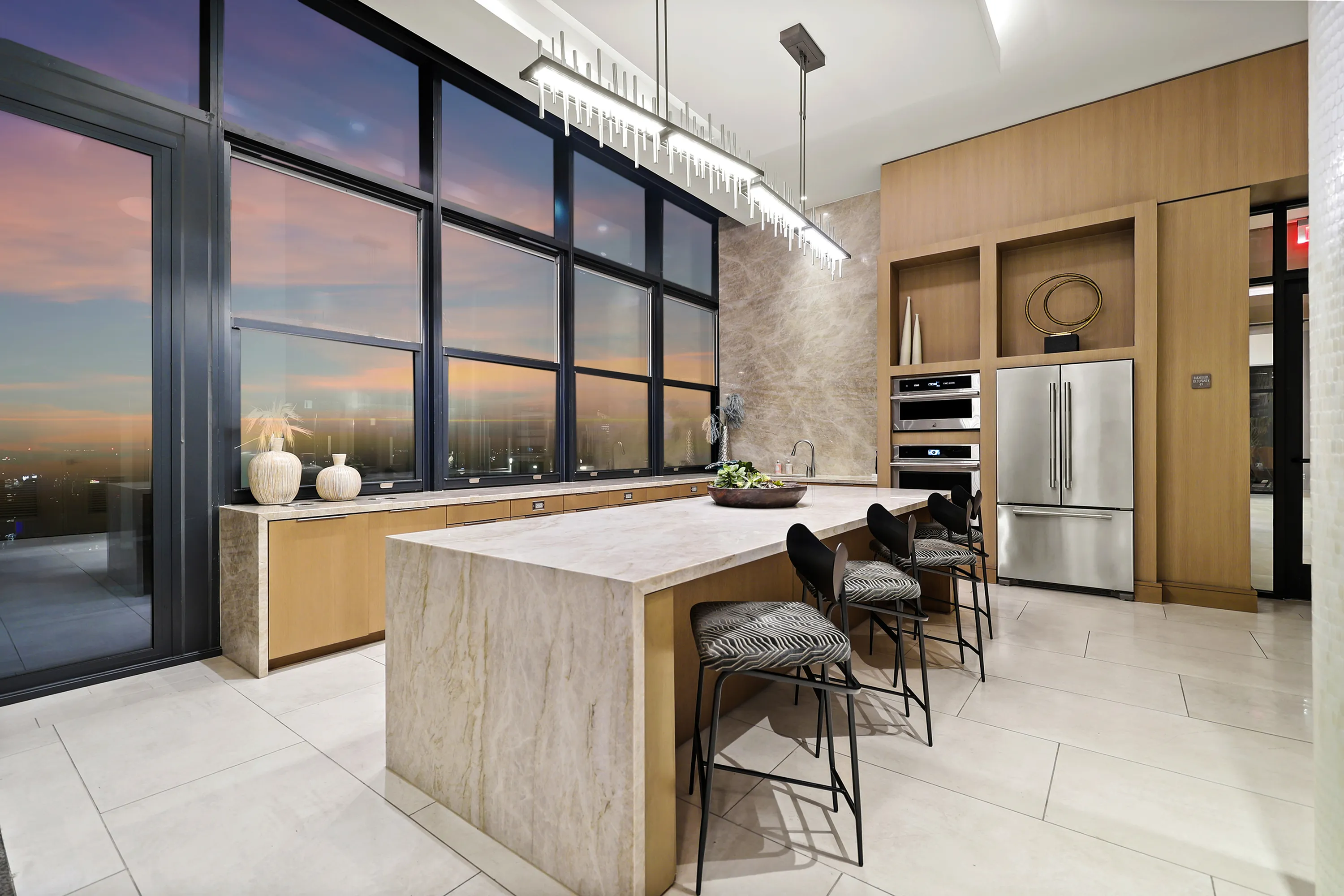 Luxurious kitchen with sleek marble countertops, stunning floor-to-ceiling windows overlooking a sunset, and modern stainless steel appliances.