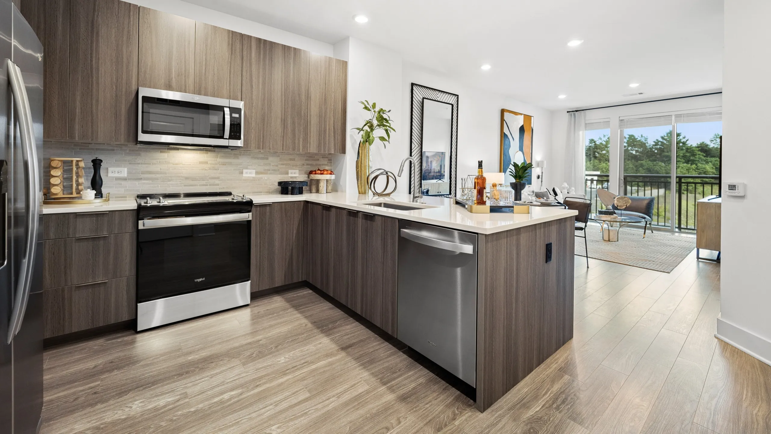 apartment kitchen with stainless steel appliances, stone counters and brown custom cabinetry with open concept living area and floor to ceiling windows