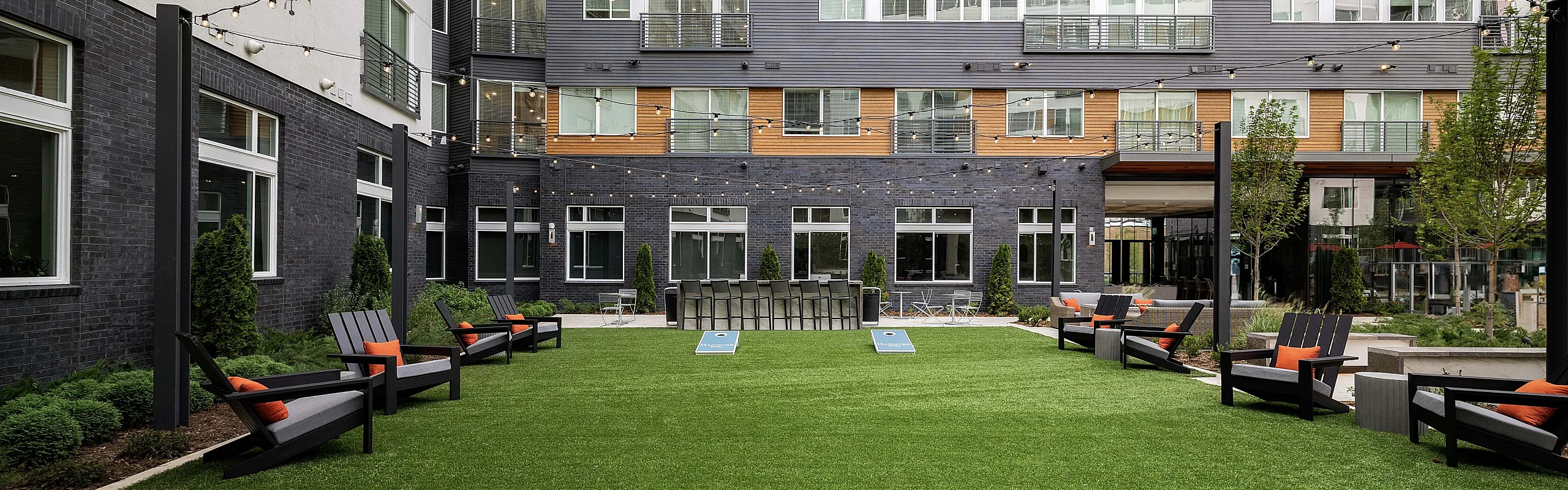Modern apartment courtyard with lush green lawn, outdoor seating, string lights, and cornhole game setup.