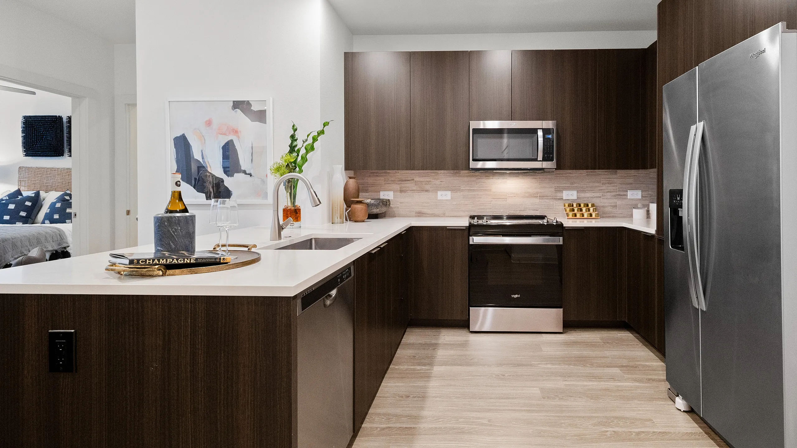 Apartment kitchen with wood-style cabinetry, stainless steel appliances, and an open layout connected to a living room and bedroom.