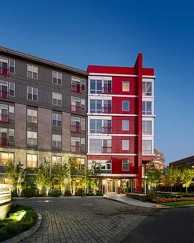 Modern apartment building with bold red accents, illuminated at dusk, surrounded by lush landscaping and a welcoming entrance.