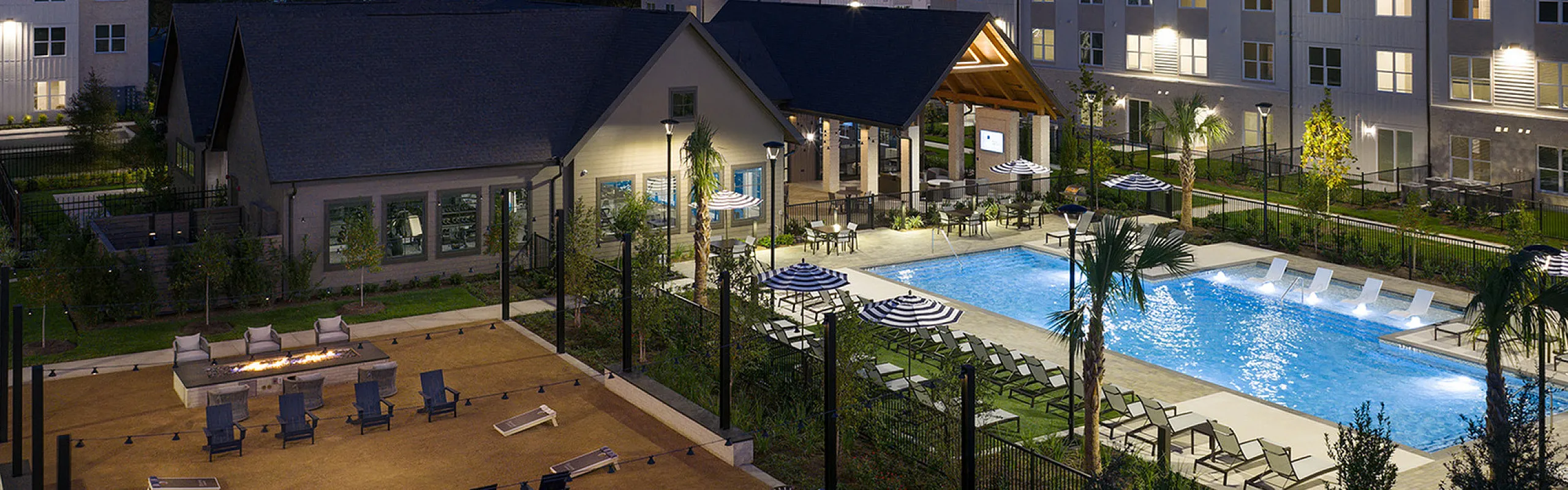Luxurious apartment complex at night featuring an illuminated swimming pool, spacious patio with lounge chairs, and a modern clubhouse for ultimate relaxation.