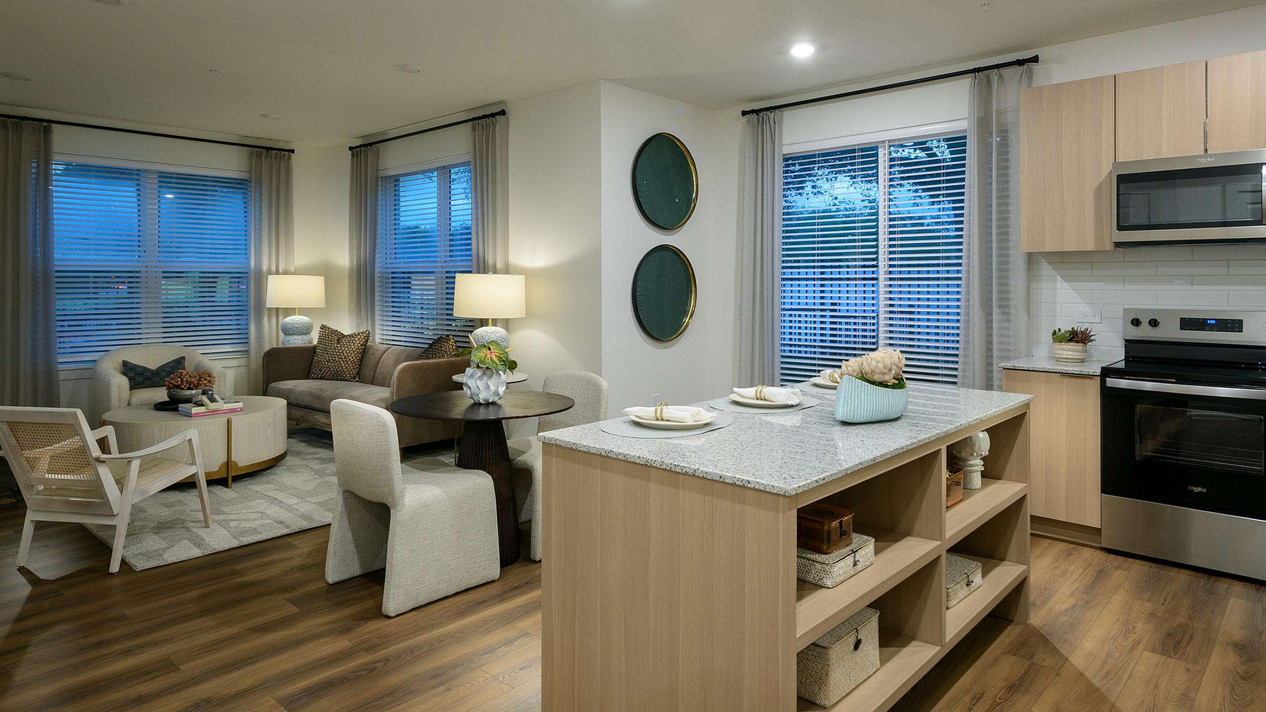 Contemporary open-plan kitchen and living room with sleek wooden flooring, cozy seating area, and modern appliances. Bright windows enhance the inviting and sophisticated ambiance.