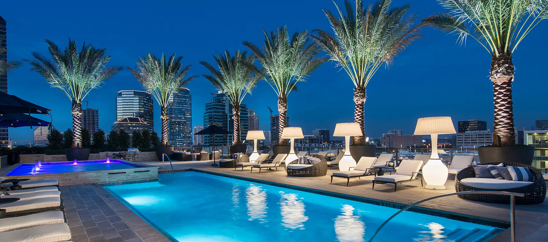 Luxurious rooftop oasis featuring an illuminated pool surrounded by elegant palm trees and stylish lounge seating, offering stunning city skyline views.