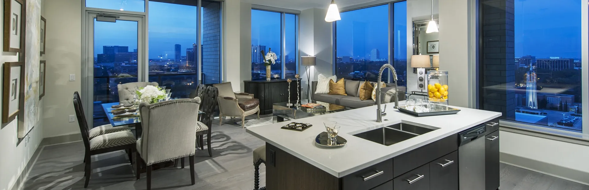 Luxurious open-concept apartment with sleek kitchen island, modern dining area, and panoramic city views at dusk.