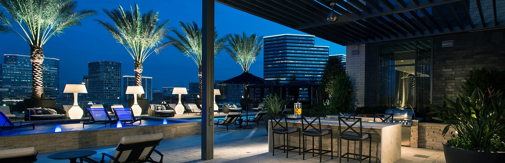 Luxurious rooftop patio at night featuring modern lounge chairs, illuminated palm trees, and stunning cityscape views.