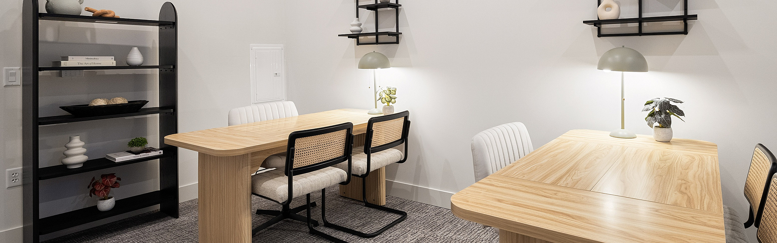 Modern workspace with two wood tables, stylish chairs, and decorative wall shelves in a cozy, well-lit room.