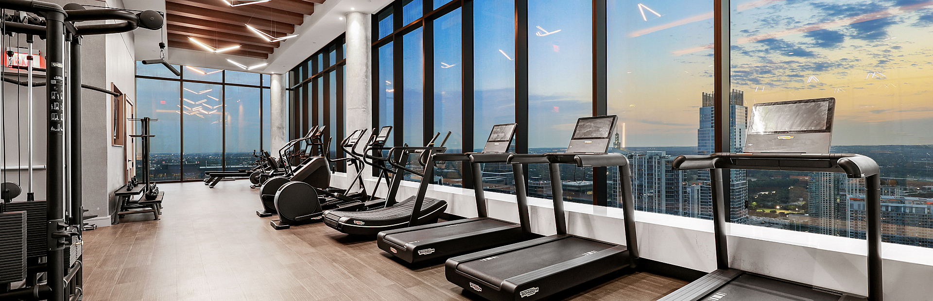 Luxurious high-rise gym with modern equipment, floor-to-ceiling windows, and panoramic city views at sunset.