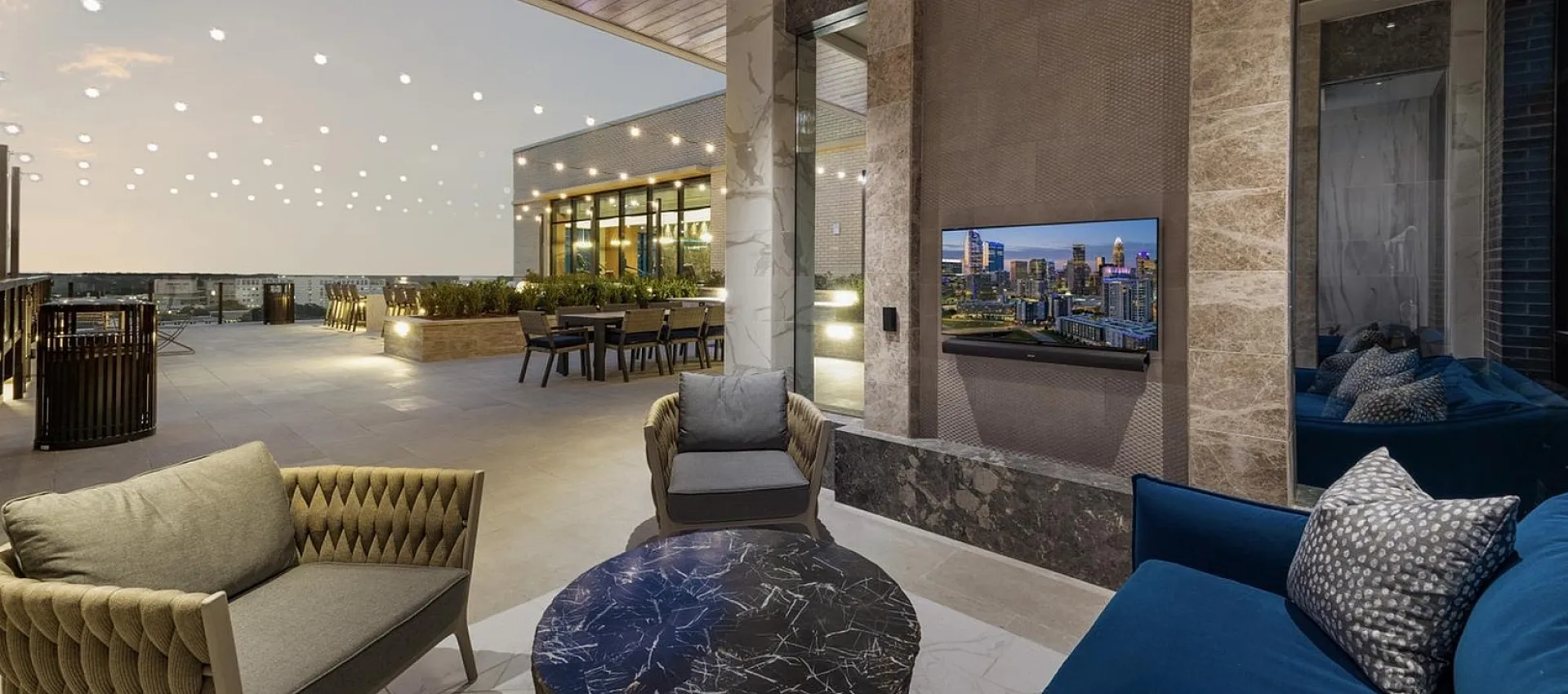 Luxurious rooftop space featuring elegant lounge chairs, stylish marble coffee table, and stunning cityscape views under dazzling string lights.