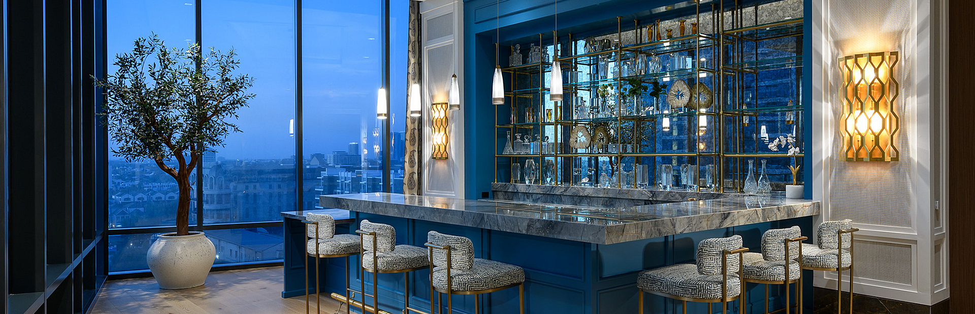 Elegant apartment bar area with sleek blue paneling, modern pendant lighting, and panoramic city views from expansive windows.