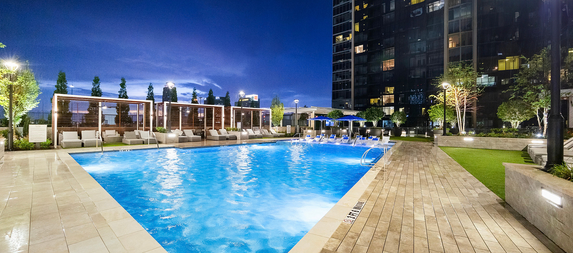 Luxurious outdoor pool area with elegant poolside seating, beautifully lit at dusk, surrounded by greenery and a modern apartment building.