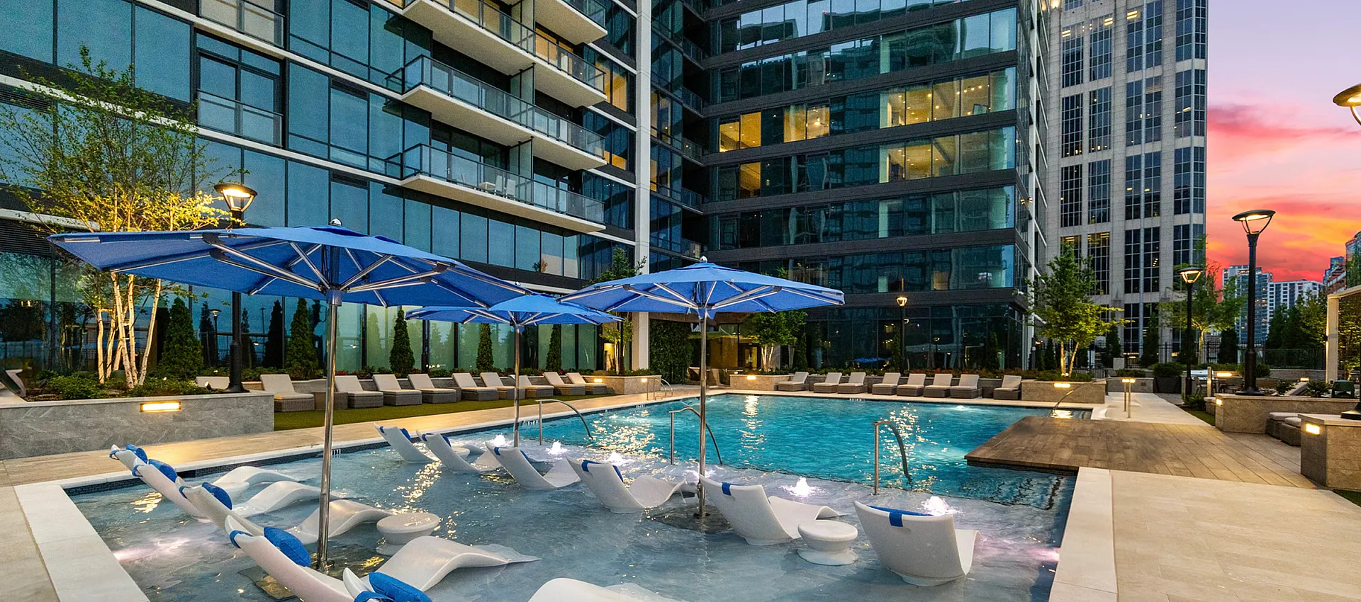 Luxurious apartment complex featuring a stunning outdoor pool with stylish lounge chairs and umbrellas against a dramatic sunset backdrop.