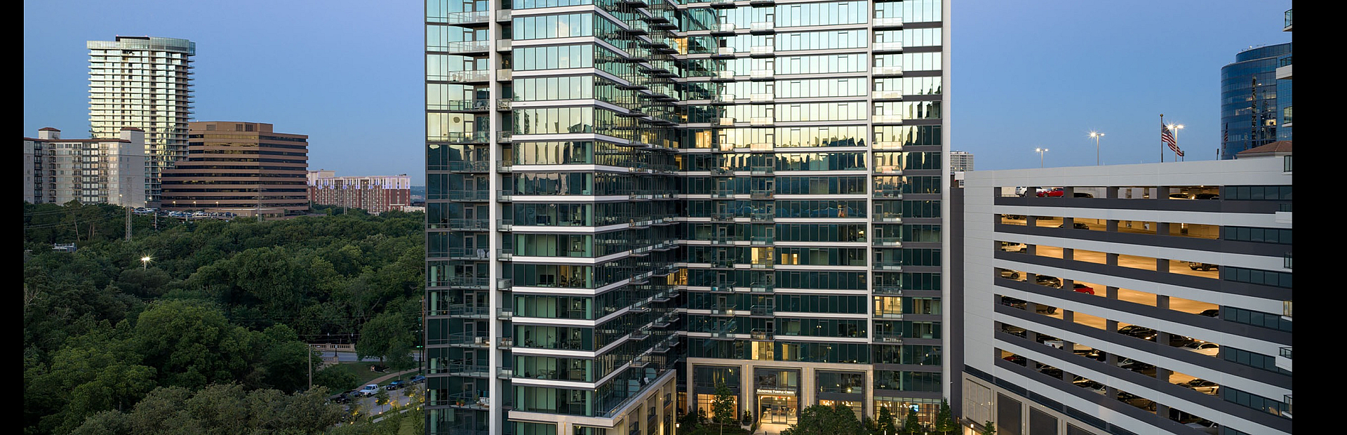 Elegant high-rise apartment building with modern glass facade, surrounded by lush greenery and urban skyline at dusk.