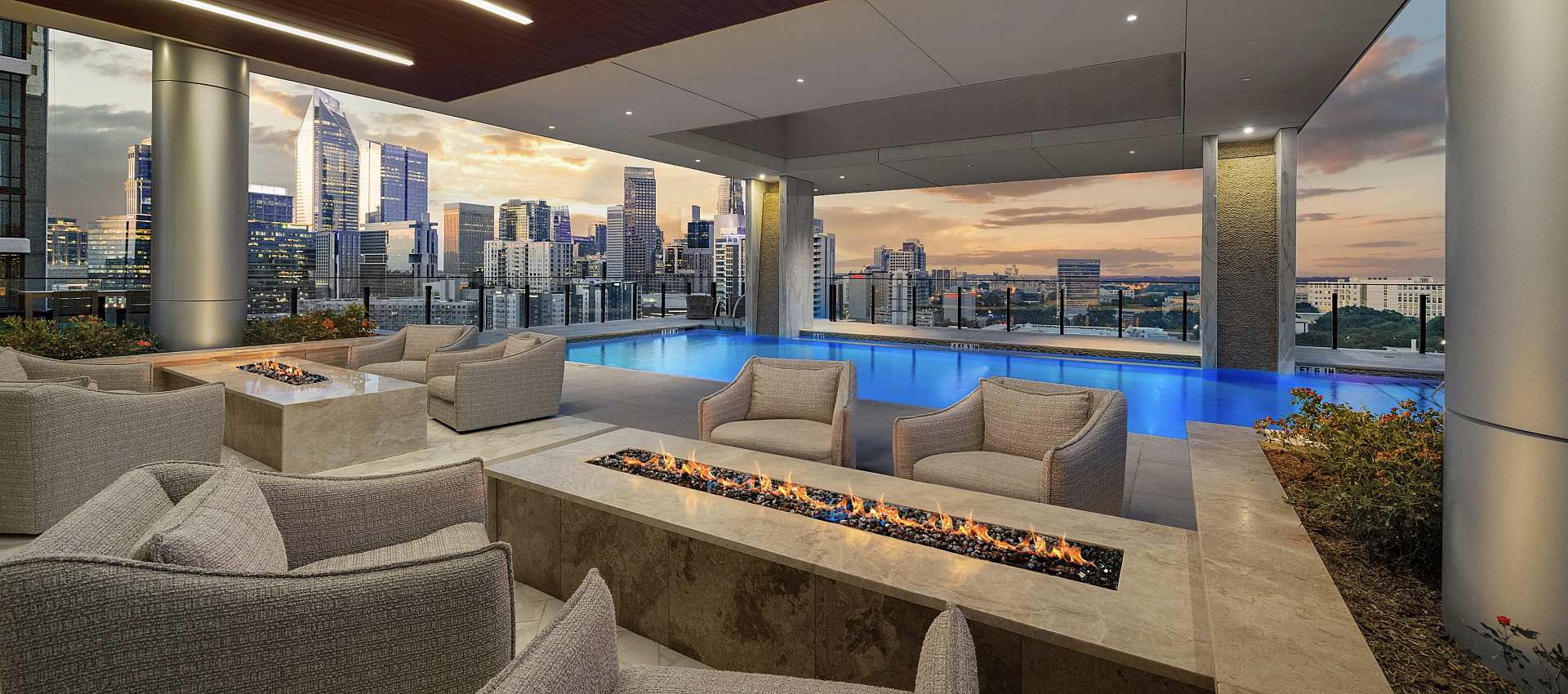 Luxurious rooftop lounge with elegant seating, a sleek modern fire pit, and a stunning pool overlooking a vibrant city skyline at sunset.