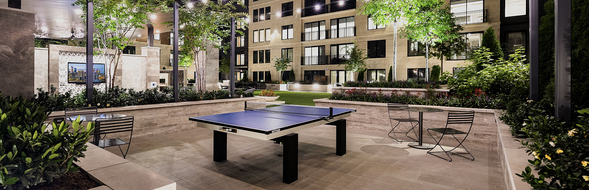 Elegant modern courtyard at night featuring a ping pong table and string lights, surrounded by lush greenery and a contemporary apartment building.