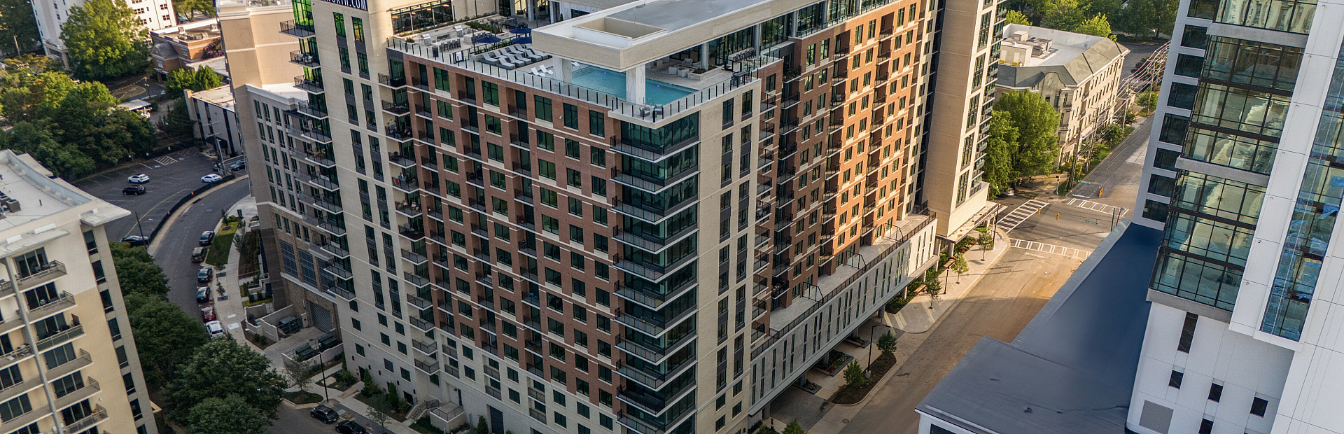 Aerial view of a modern, luxurious high-rise apartment building with a rooftop pool, nestled in a vibrant urban neighborhood surrounded by lush greenery.