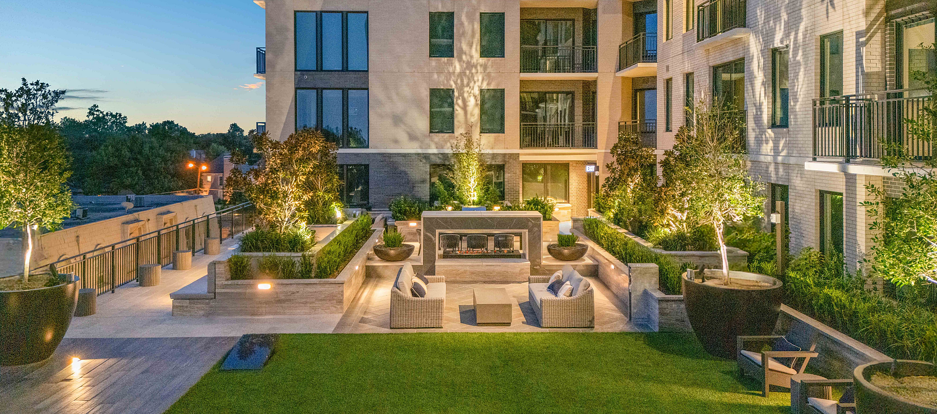 Luxurious apartment building at twilight with modern balconies and a beautifully lit courtyard featuring elegant seating and lush greenery.