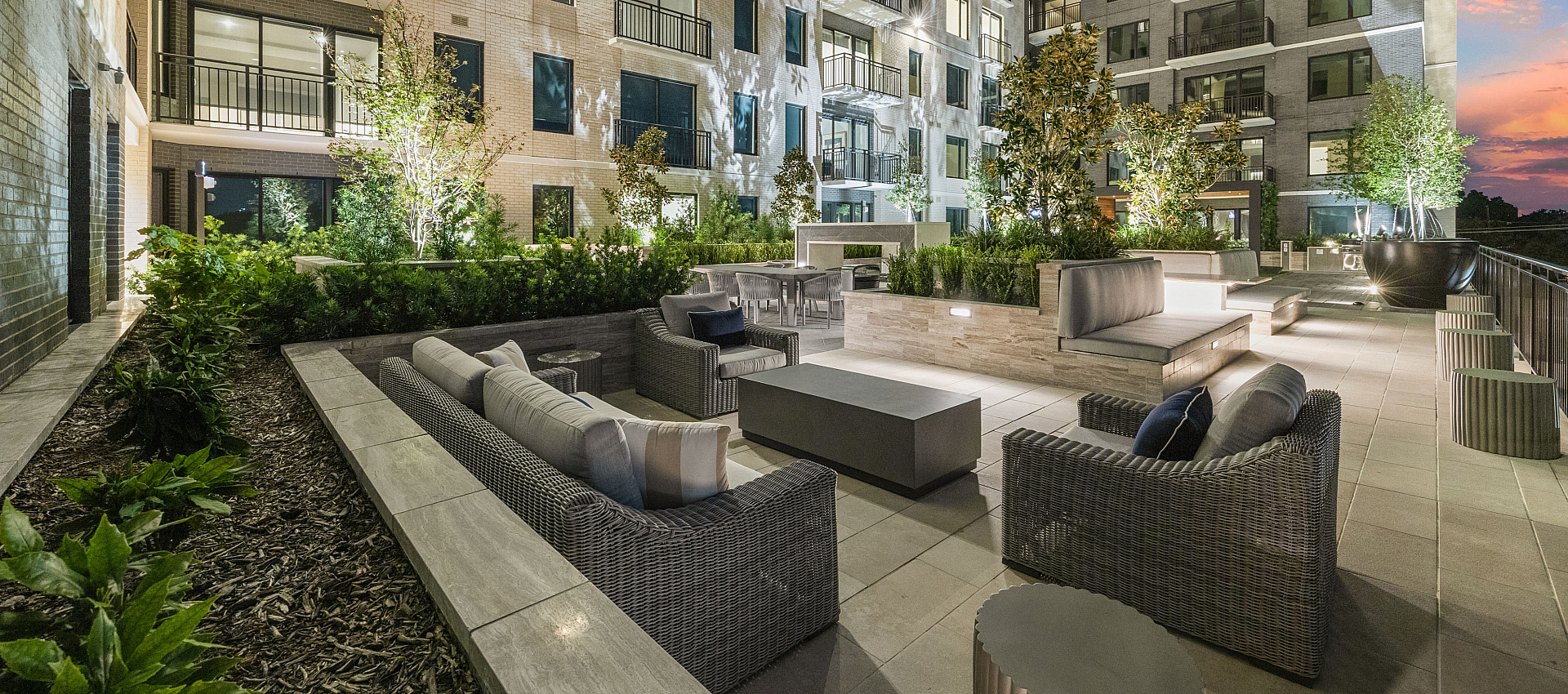 Modern apartment exterior showcasing a beautifully lit evening courtyard with elegant patio furniture and lush greenery.
