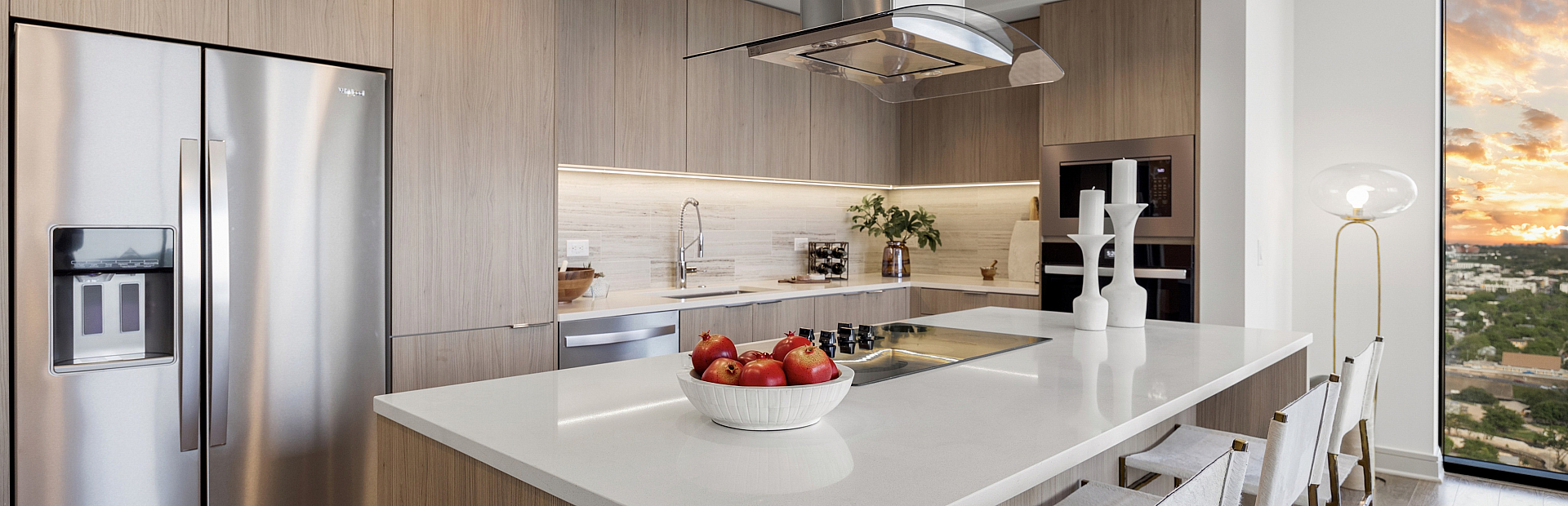 Modern kitchen with sleek wooden cabinetry, stainless steel appliances, and an elegant island boasting a bowl of fresh apples, overlooking a stunning cityscape at sunset.