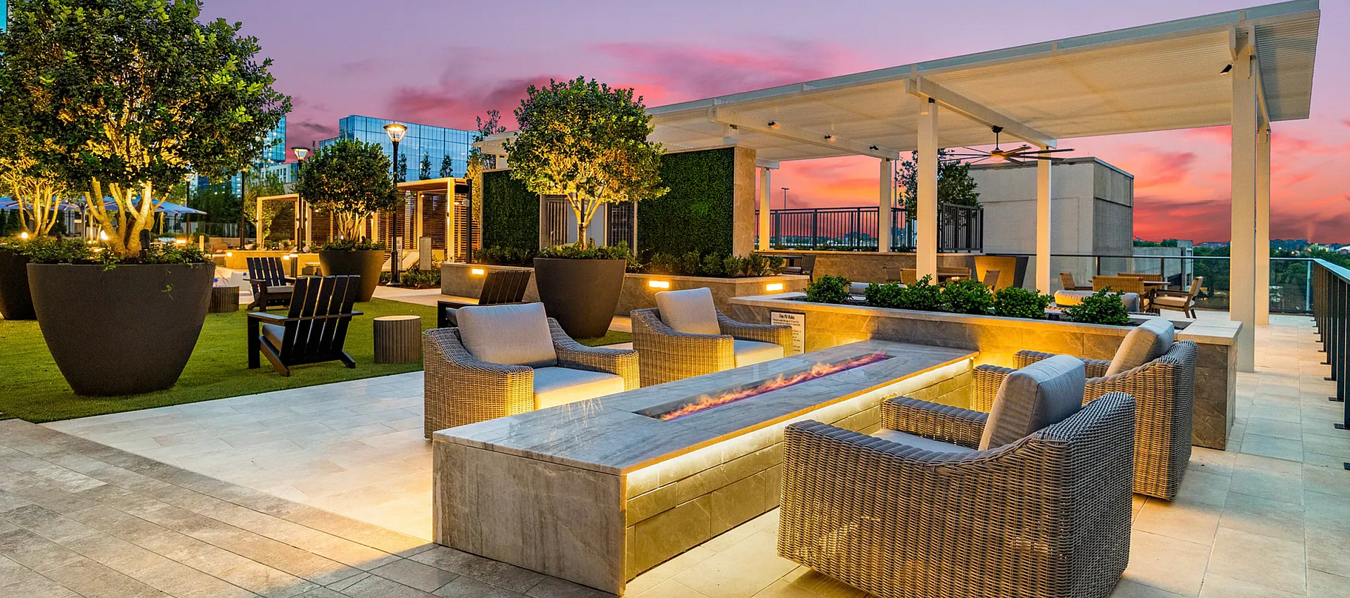 Luxurious rooftop lounge with modern wicker seating, ambient lighting, and lush greenery against a stunning sunset backdrop.