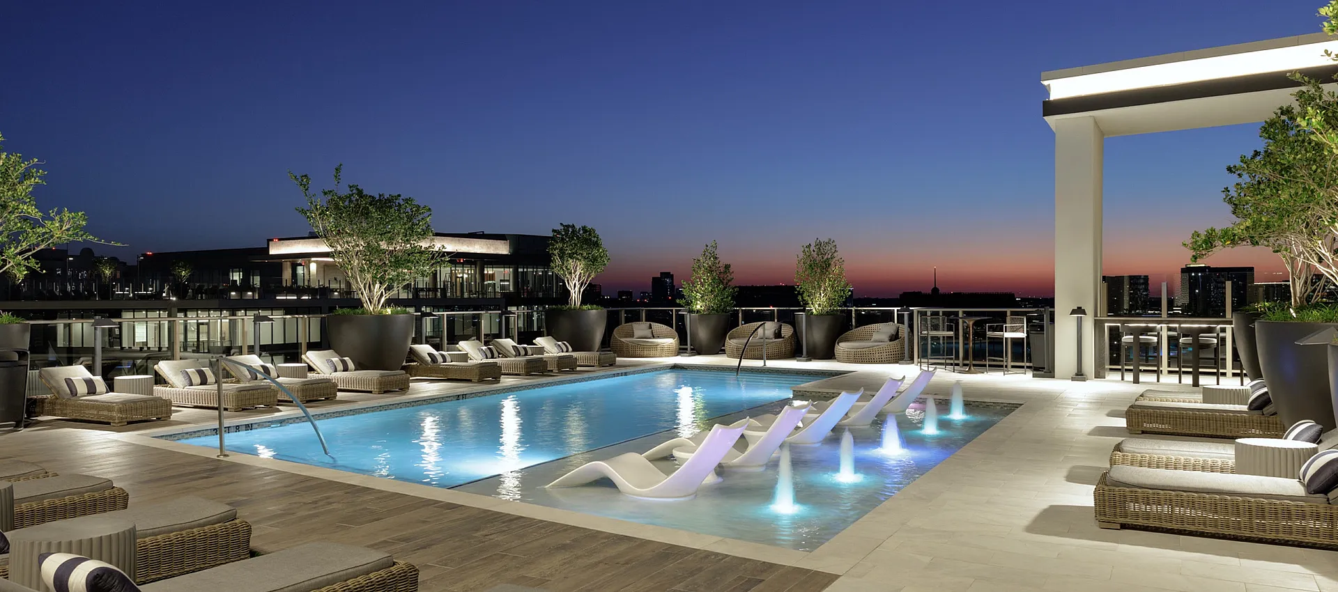 Luxurious rooftop pool area at sunset featuring elegant lounge chairs, illuminated fountains, and panoramic city views.