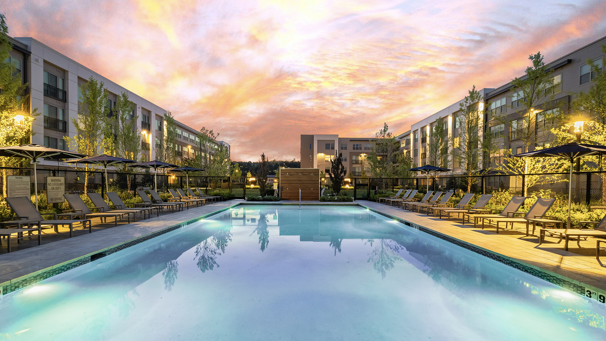 Luxurious apartment complex featuring a stunning outdoor pool, surrounded by elegant lounge chairs and lush greenery, set against a breathtaking sunset sky.