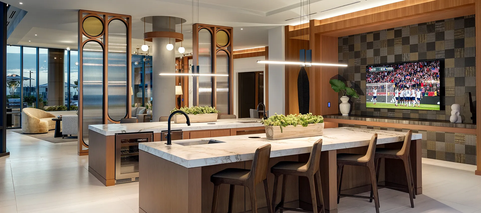 Luxurious open-concept kitchen with modern decor, sleek marble countertops, and stylish lighting fixtures, featuring a cozy living area with a large TV.