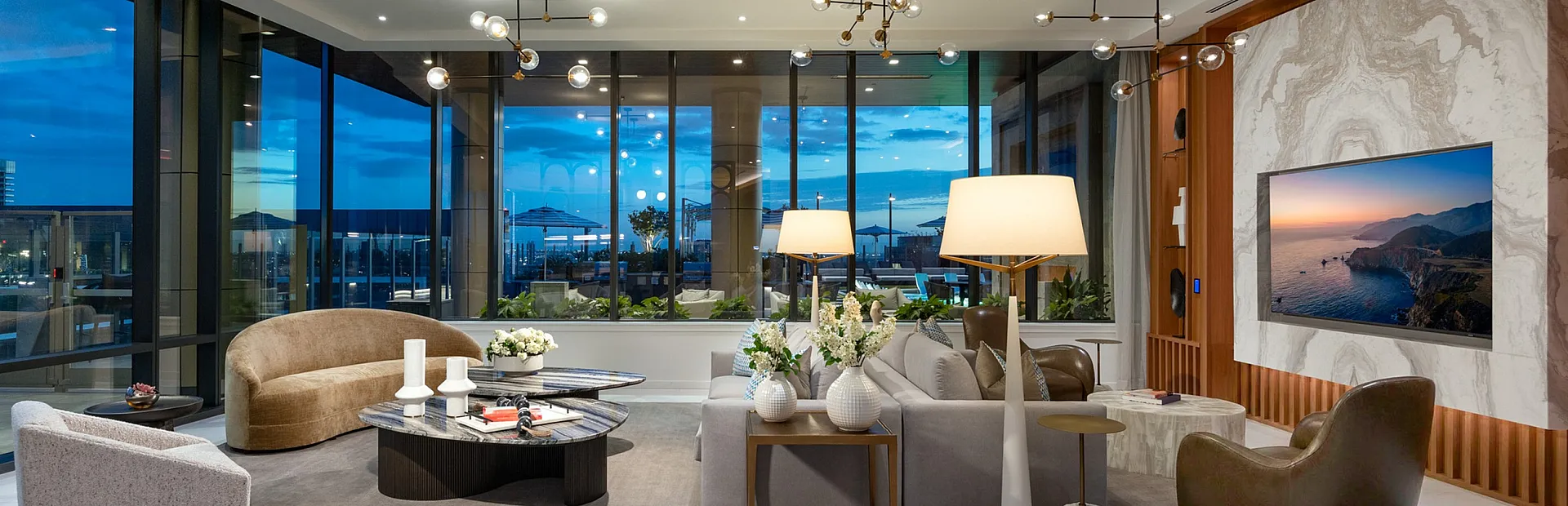 Luxurious living room with modern lighting, plush seating, and expansive floor-to-ceiling windows offering breathtaking city views at dusk.