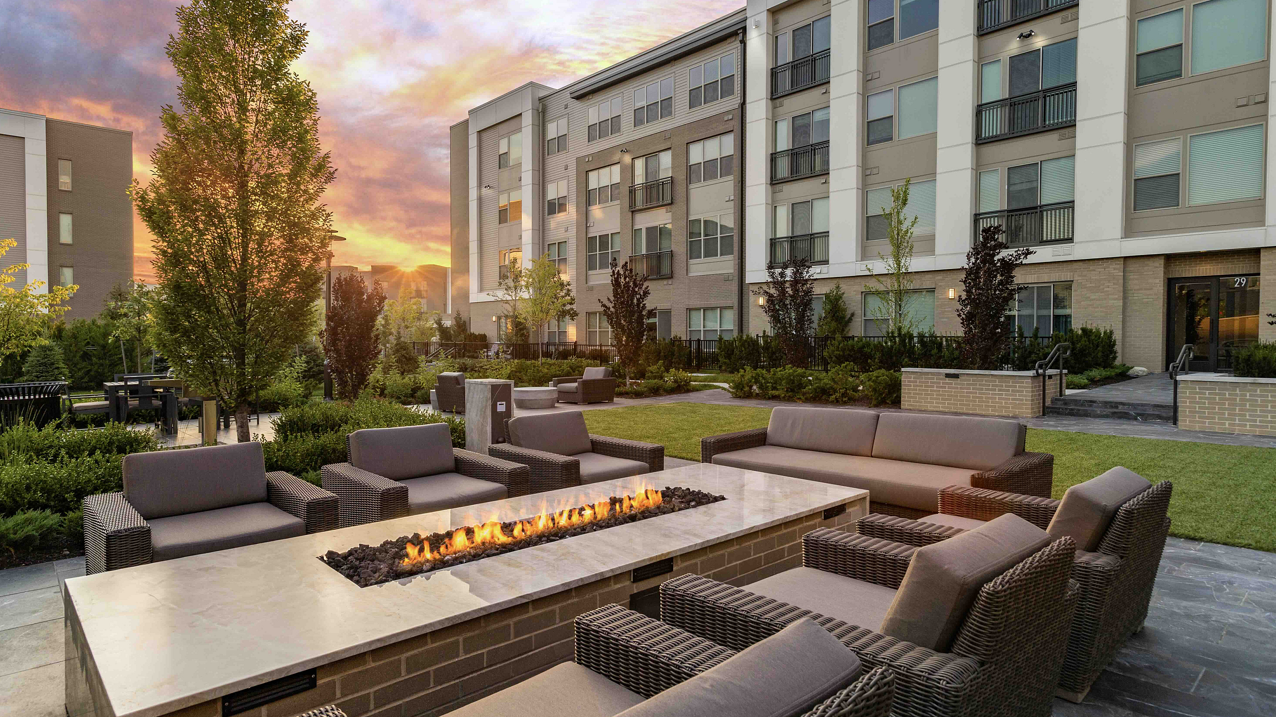 Chic outdoor lounge area with modern fire pit, surrounded by lush landscaping and contemporary apartment building at sunset.