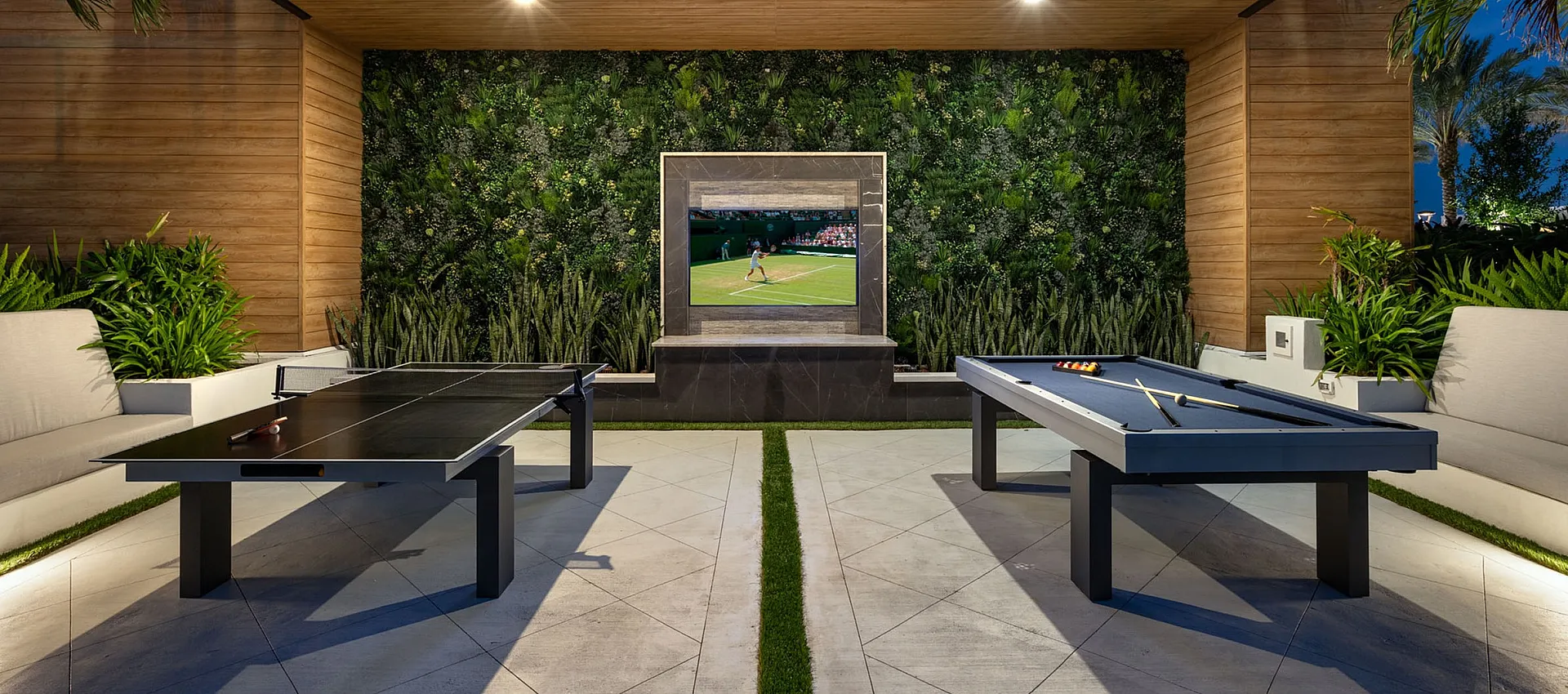 Modern outdoor entertainment area with stylish pool and ping pong tables, lush greenery, and a wall-mounted TV, perfect for evening gatherings.