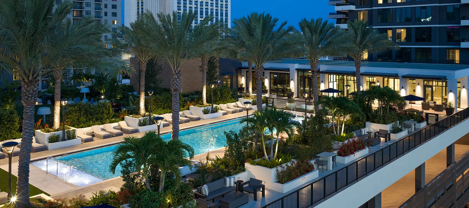 Luxurious apartment complex at dusk featuring a stunning rooftop pool surrounded by elegant palm trees and modern architecture.
