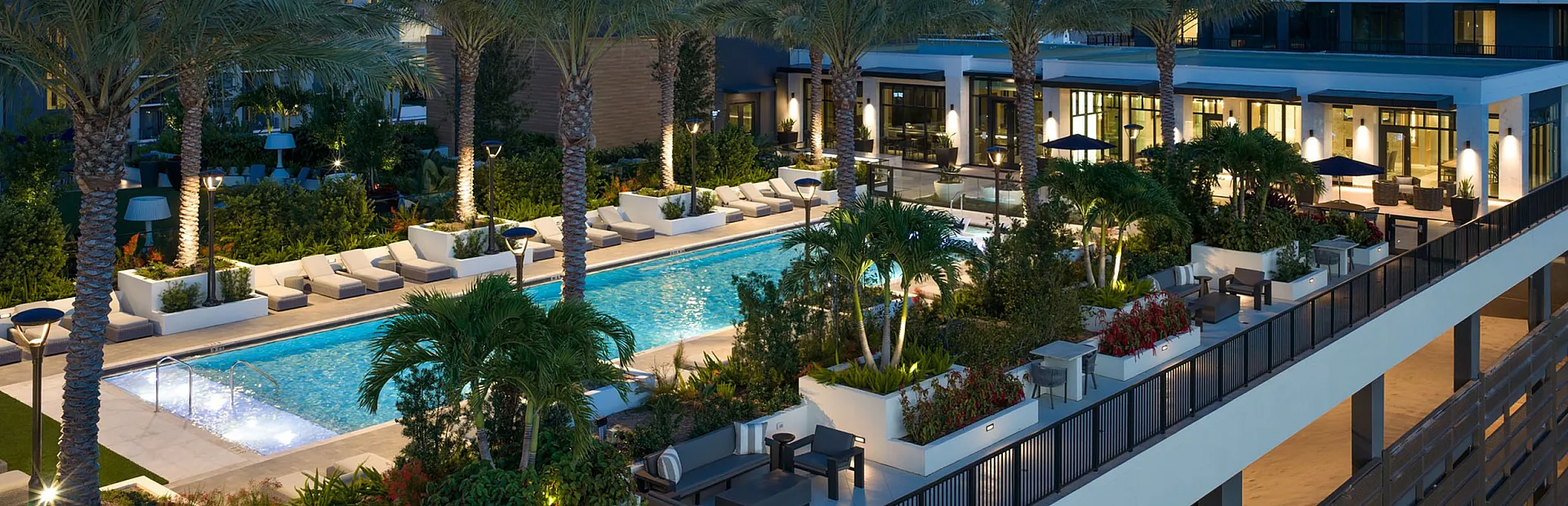 Luxurious apartment complex featuring a beautifully lit outdoor pool area, surrounded by palm trees and modern high-rise buildings in an urban setting.