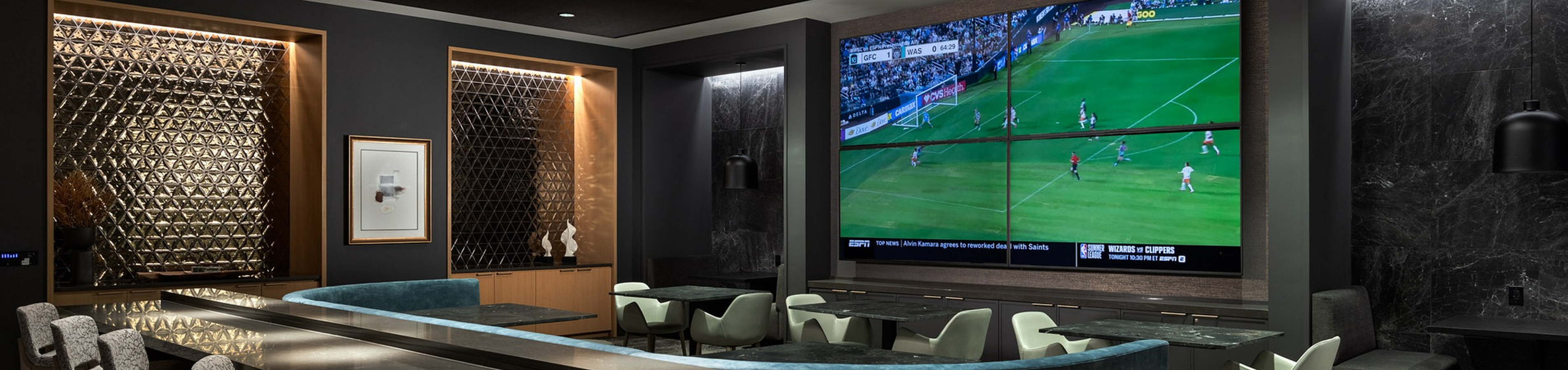 Luxurious recreation area featuring a sleek bar with modern seating, elegant wall decor, and a large screen displaying a sports match for ultimate entertainment.