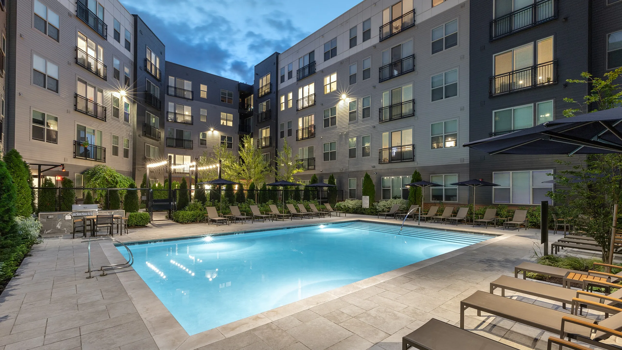 Modern apartment complex with a stunning illuminated pool area, surrounded by comfortable lounge chairs and lush greenery.