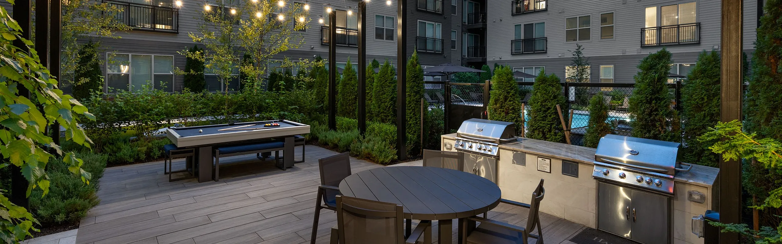 Chic outdoor patio with modern seating, ambient string lights, grills, and vibrant greenery, set against a stylish apartment facade.