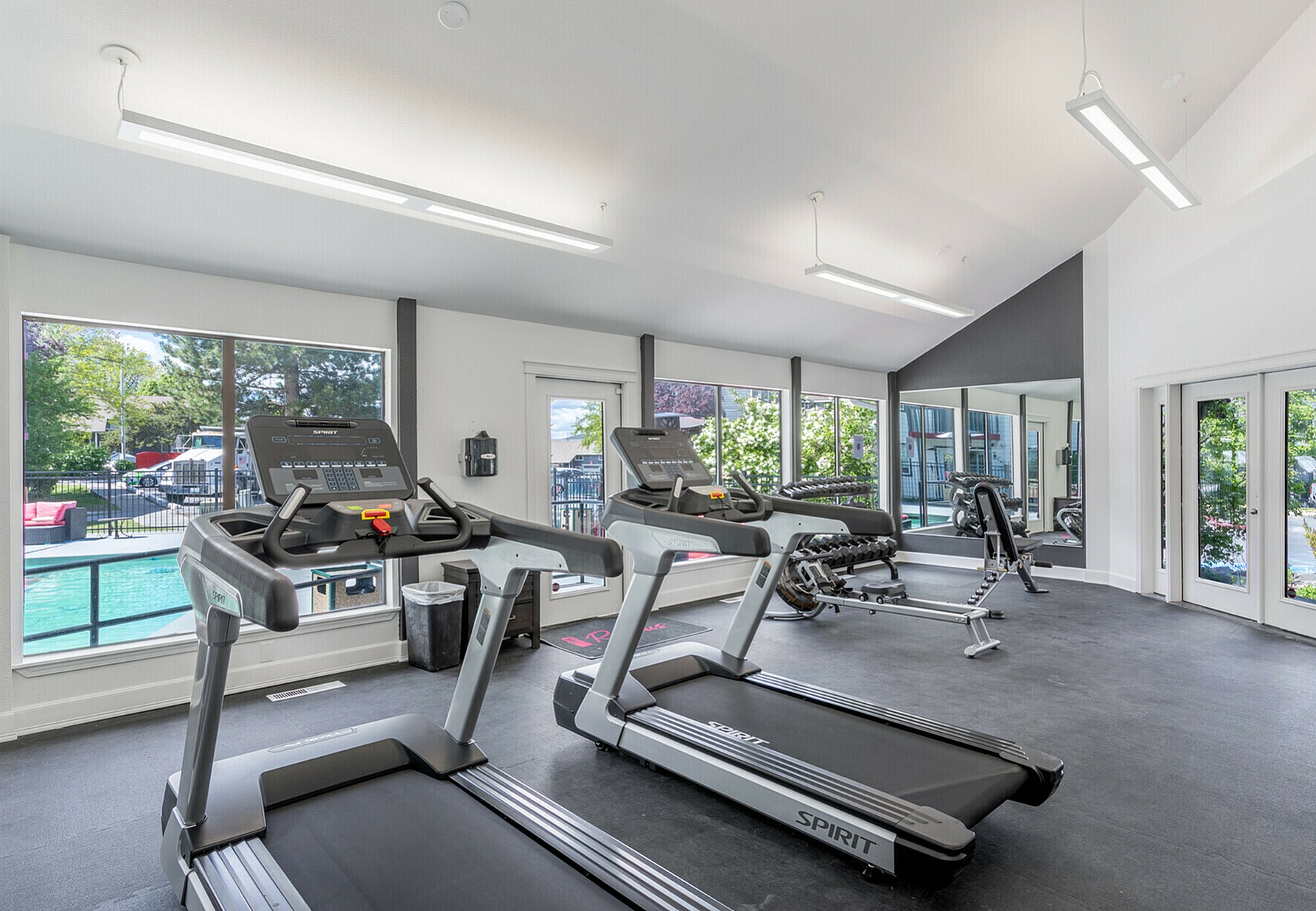 Bright and spacious apartment gym with modern fitness equipment and large windows overlooking a pool.
