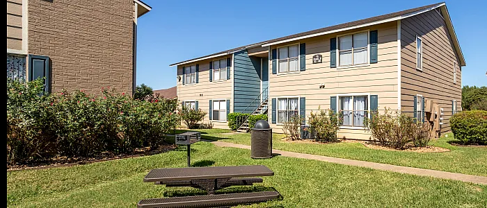 Charming two-story apartment buildings with lush green lawns, inviting paths, vibrant shrubbery, and a welcoming picnic area under a clear blue sky.
