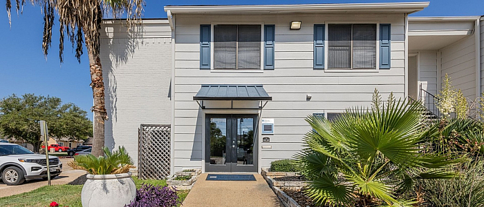 Charming apartment building entrance with lush landscaping and modern exterior, showcasing vibrant plants and a welcoming pathway under a clear blue sky.