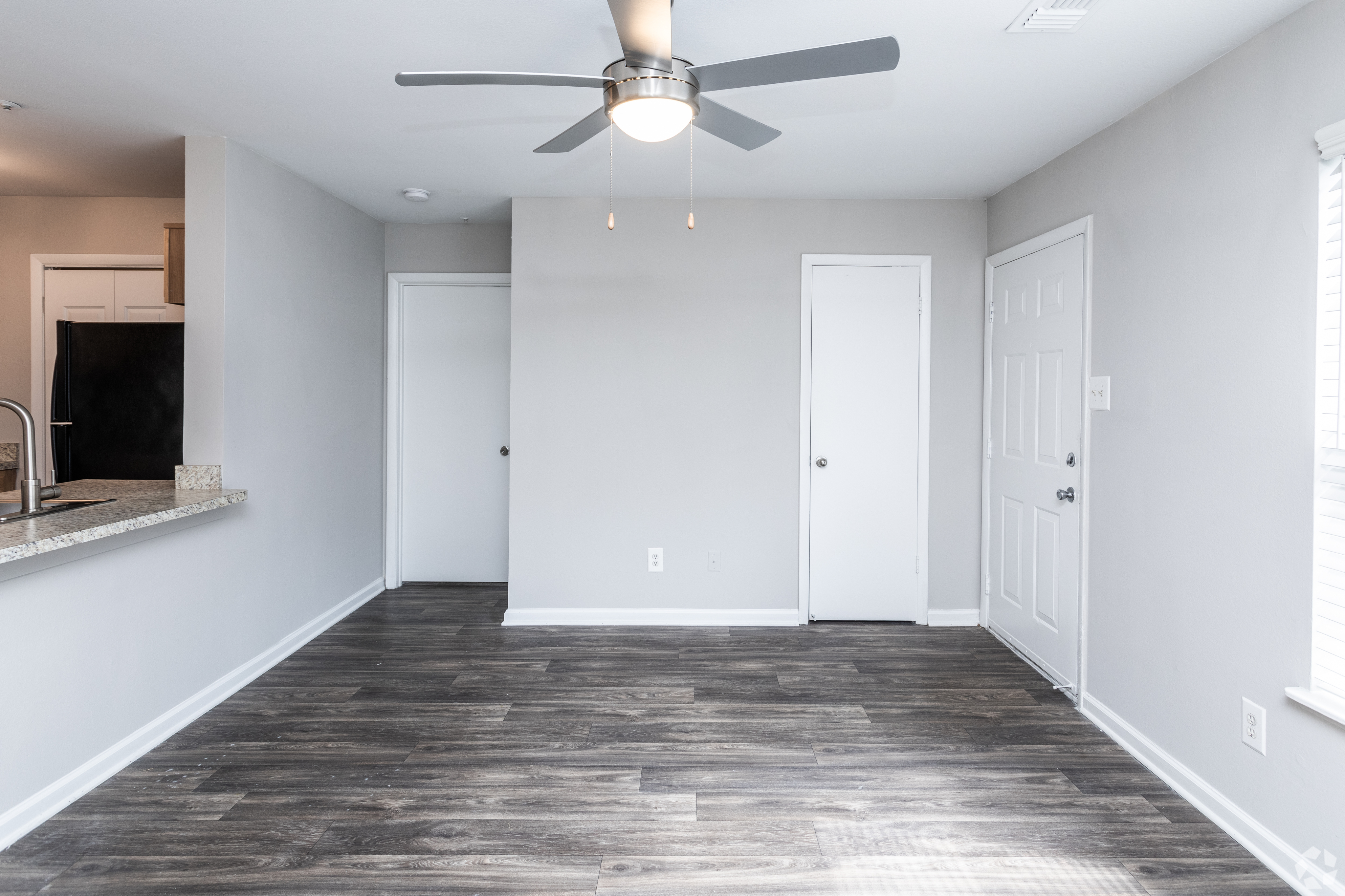 Modern living room with sleek wood flooring and a ceiling fan, adjacent to a kitchen area.