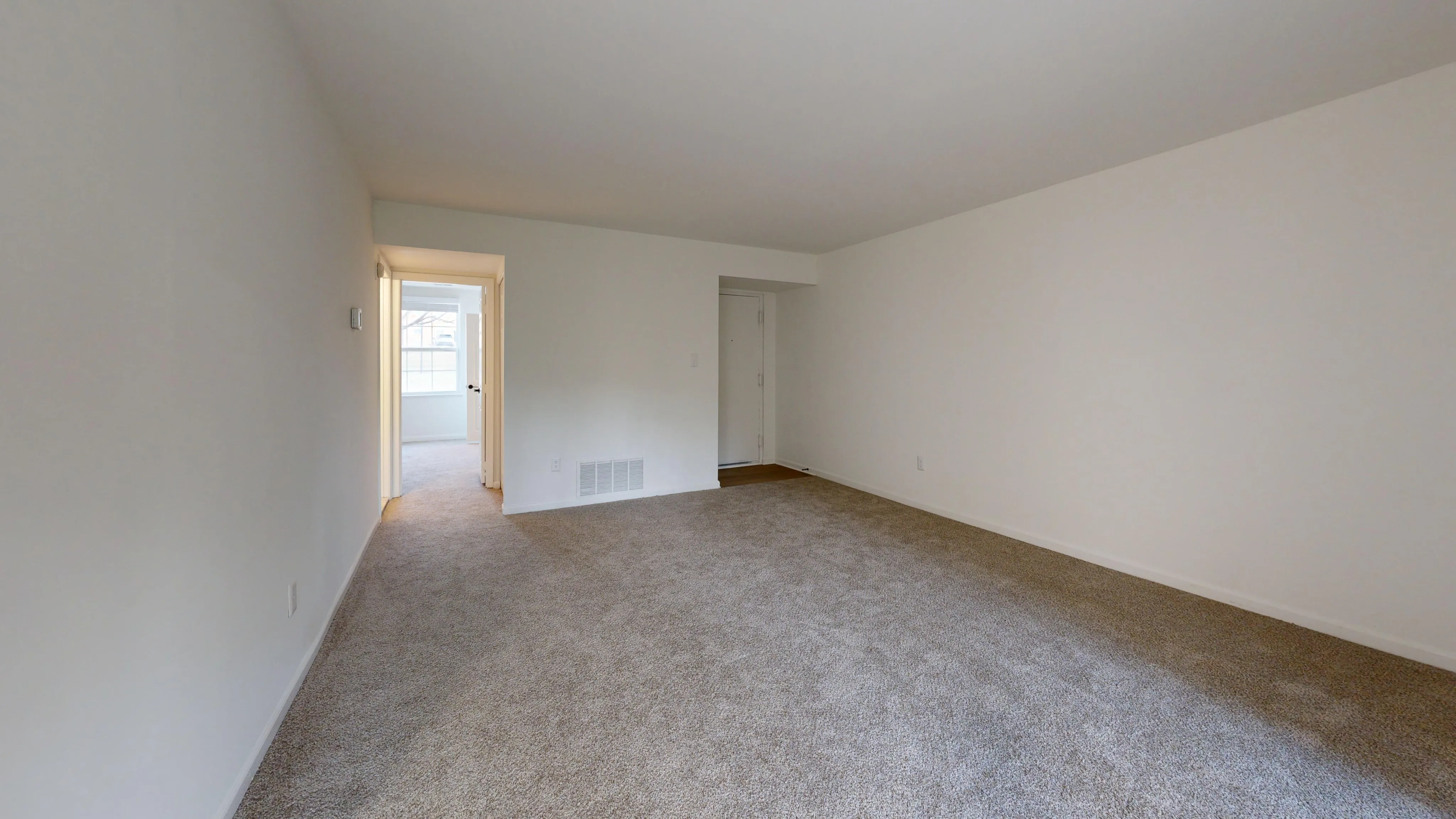 Inviting living room with plush carpet, neutral walls, and bright natural light leading to a cozy bedroom.