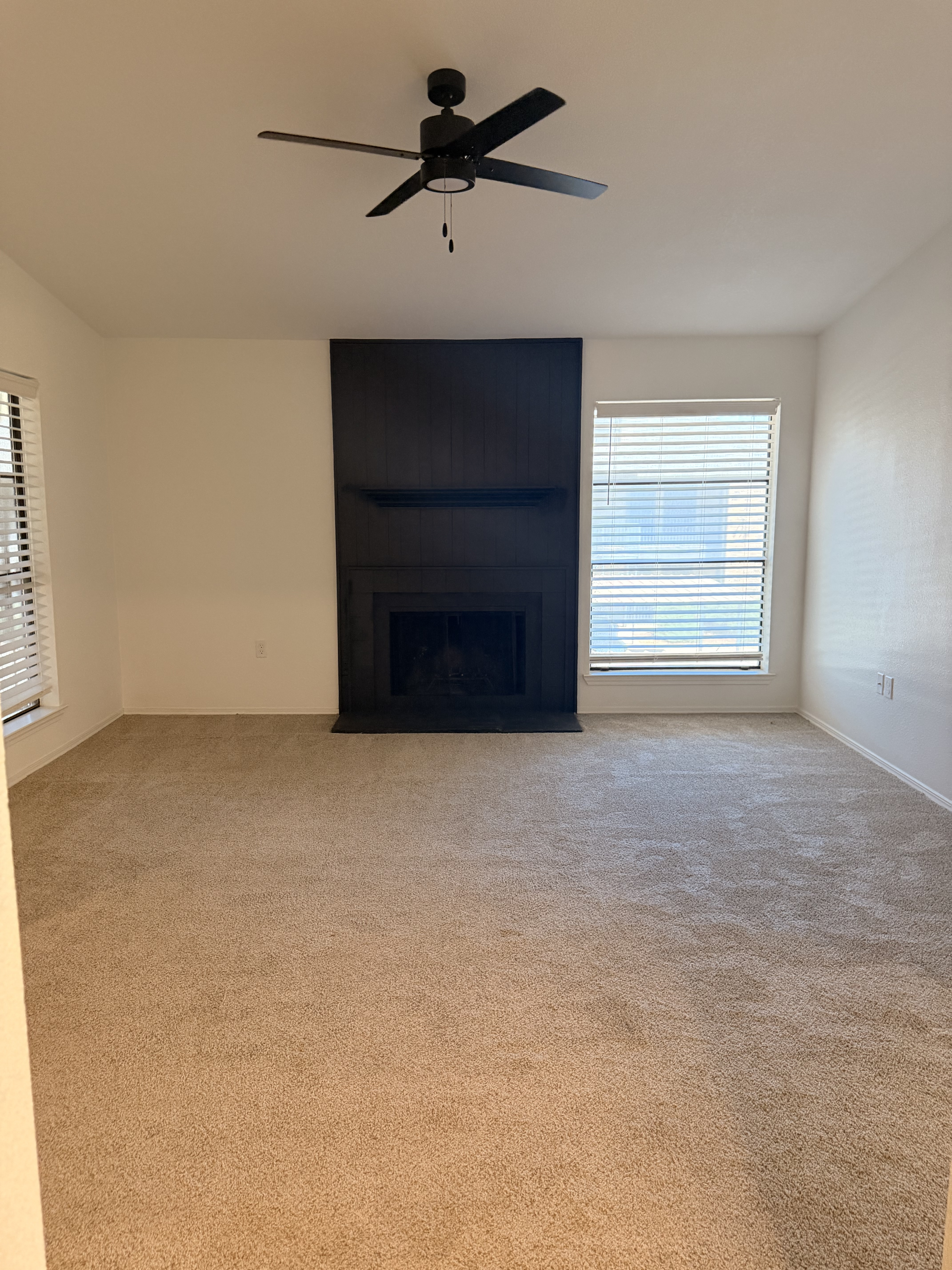 Inviting living room showcasing plush carpet, a modern ceiling fan, and a stylish black fireplace flanked by large windows.
