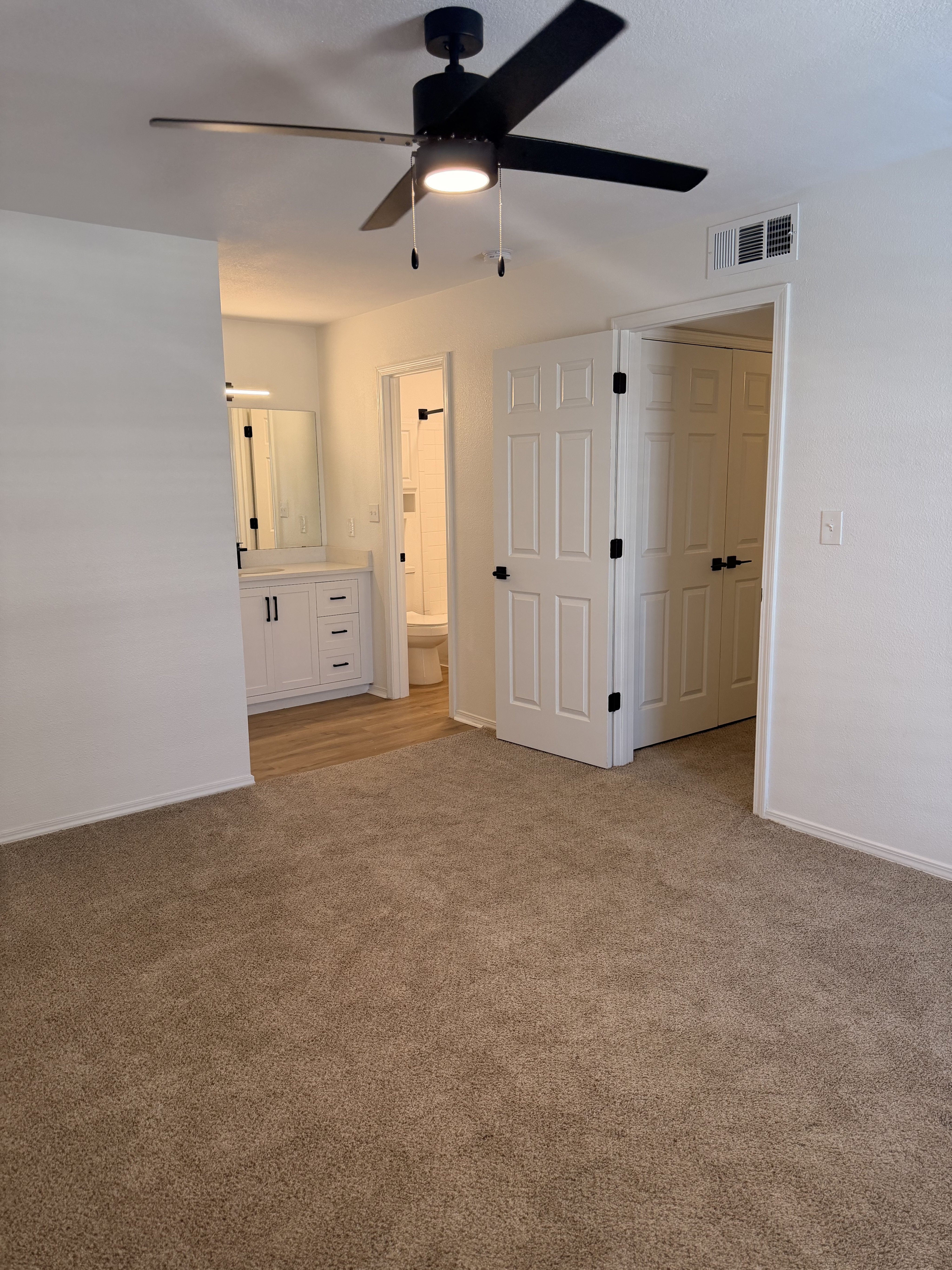 Inviting bedroom featuring plush carpet, modern ceiling fan, and an en-suite bathroom with vanity in a stylish apartment.