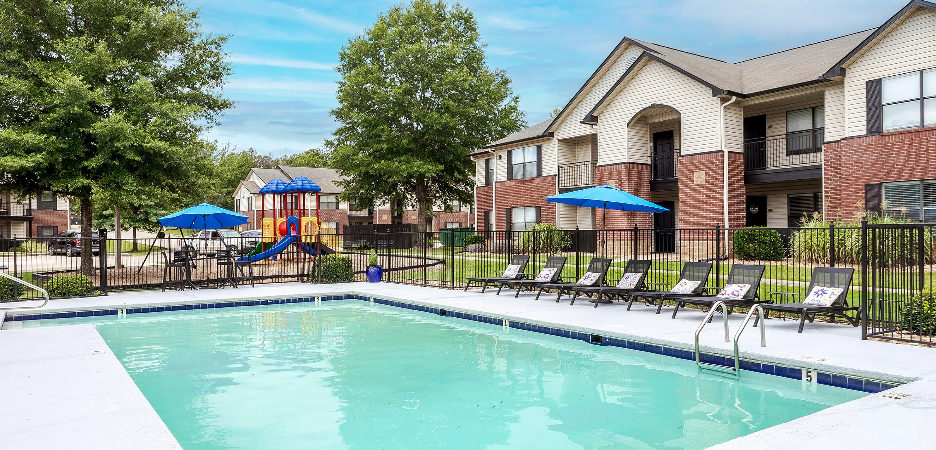 Inviting apartment community pool with lounge chairs and a children&rsquo;s playground in the background.