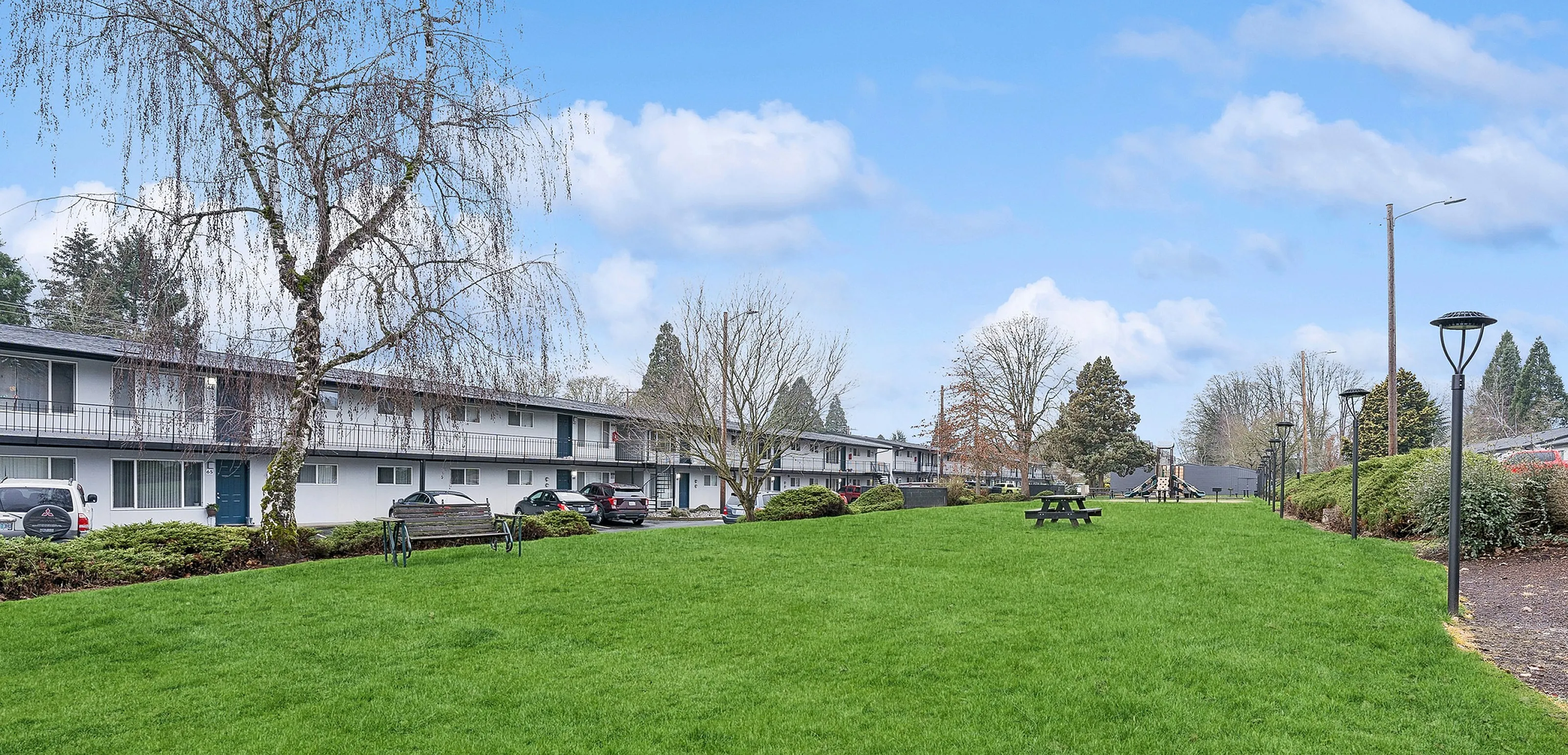 Inviting apartment complex with lush green lawns, outdoor benches, and serene landscaping under a clear blue sky.