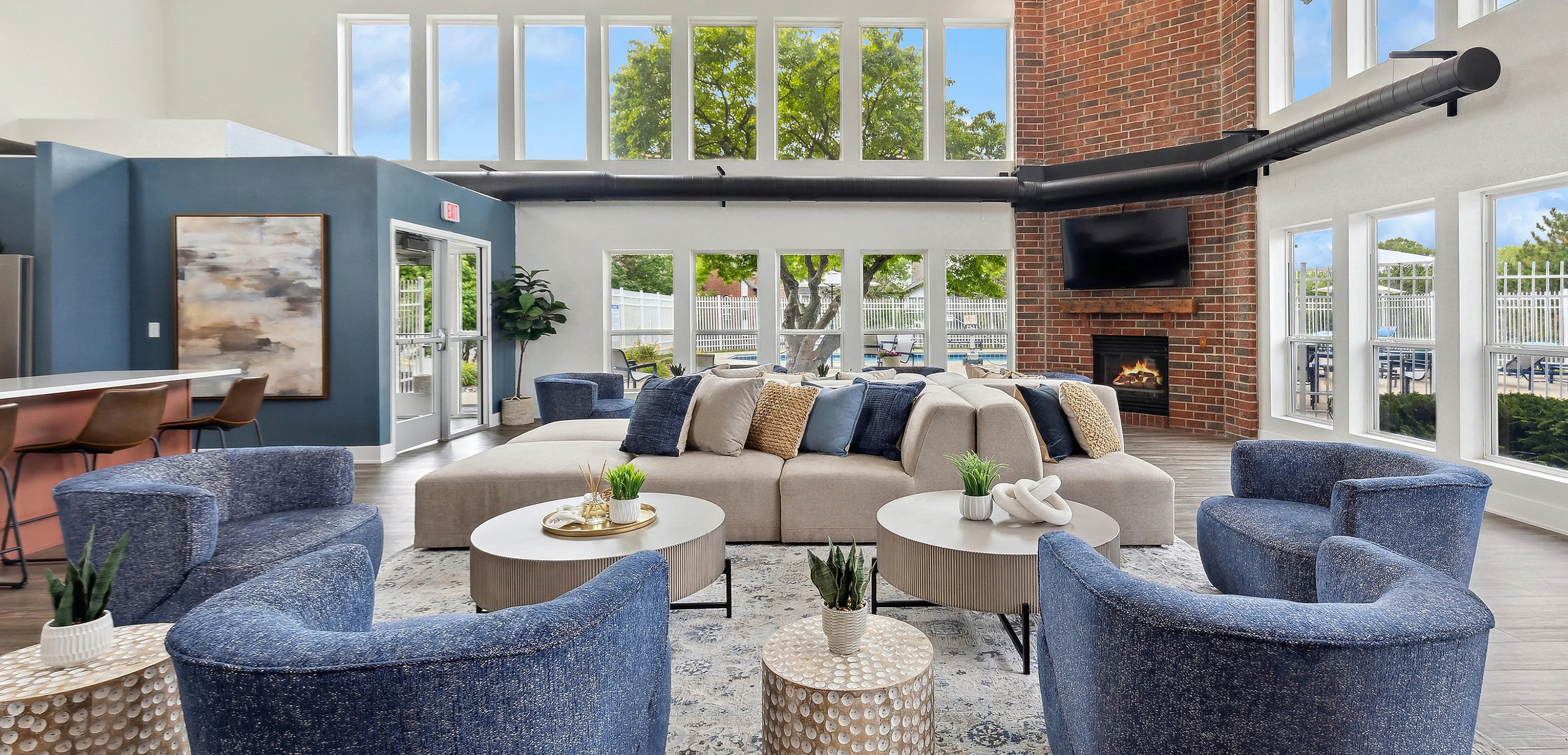 Bright and inviting living space featuring modern furnishings, a brick accent wall, a cozy fireplace, and large windows with views of greenery.